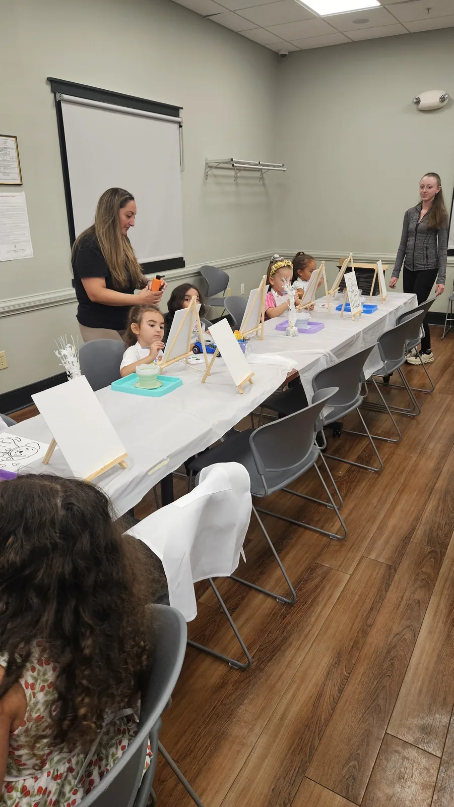 Instructor leading children at a long craft table in a decorated community room