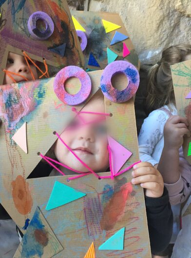 Child holding a painted cardboard monster mask with button eyes at a STEAM Sprouts craft class