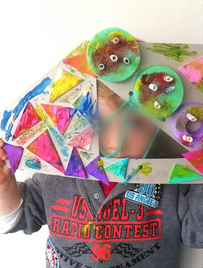 Child holding a painted cardboard monster mask with green button eyes at a STEAM Sprouts craft class