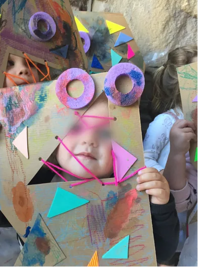 Child holding a painted cardboard monster mask with button eyes, face blurred